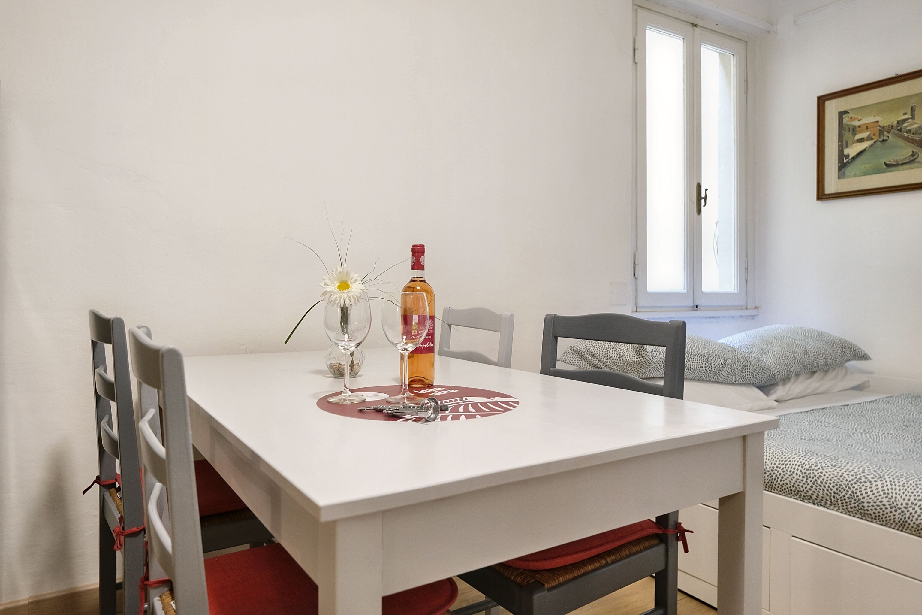 A small, neatly arranged room with a white table set for two, a single bed with patterned bedding, and a window with a view of a building.