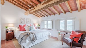 A bedroom with a wooden ceiling, a bed with white bedding, red pillows, and a chair with a red cushion.