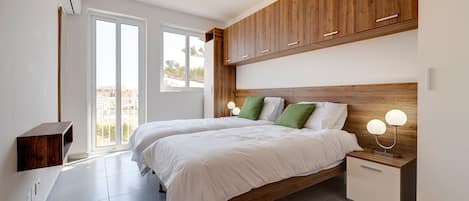 A bedroom with two beds, wooden headboard, bedside tables with lamps, and a view through the door.
