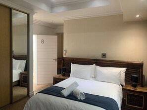 Una habitación de hotel con una cama grande, cabecera de madera, dos mesitas de noche con lámparas, un espejo y una puerta.