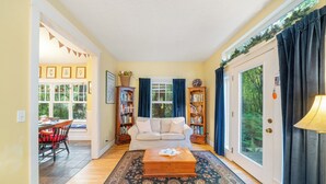 Ein Wohnzimmer mit einer Couch, einem Couchtisch und einem Teppich. Zwei Fenster mit Vorhängen, eine Tür und ein Bücherregal sind vorhanden.