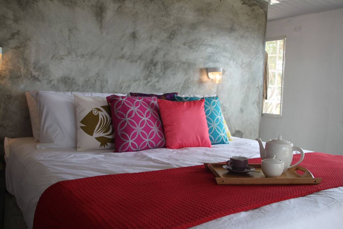 Ein Bett mit einer roten Decke, Kissen mit verschiedenen Mustern und ein Tablett mit Teekanne und Tasse.