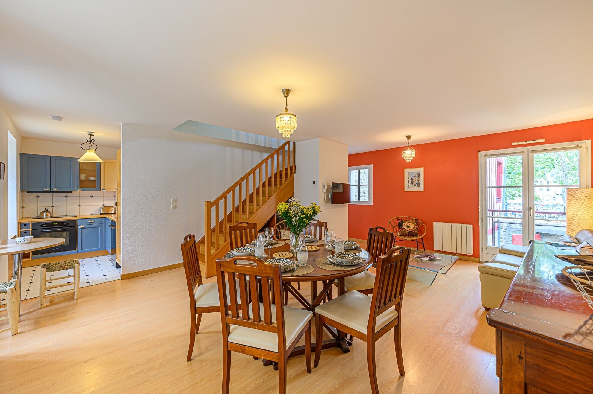 Un comedor amplio con una mesa de madera dispuesta para seis personas, rodeada de sillas. Una escalera conduce a un nivel superior.