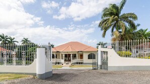 Une entrée fermée par un portail mène à une maison avec un toit recouvert de tuiles rouges, une véranda et un panneau indiquant le numéro de la maison.