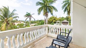 Un balcon avec une balustrade blanche, deux chaises noires et une vue sur des palmiers et un petit bâtiment au loin.