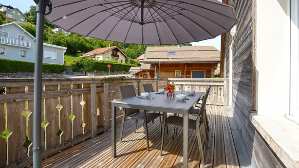 Een houten terras met een tafel, stoelen en een grote parasol, met op de achtergrond huizen.
