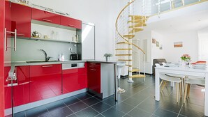 Een moderne keuken met rode kasten, een wit aanrechtblad en een vloer van zwarte tegels. Er is een zithoek met een witte tafel en stoelen, en een spiltrap die naar een hoger gelegen verdieping leidt.