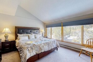 Um quarto com uma cama grande, duas luminárias de cabeceira, uma janela com vista para árvores e uma cadeira.