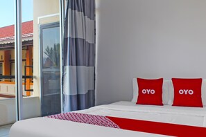 Une chambre d’hôtel avec un lit, deux oreillers rouges ornés du mot « OYO », un plaid rouge et blanc à motifs, et une vue sur un immeuble avec un toit rouge, visible à travers une porte vitrée.