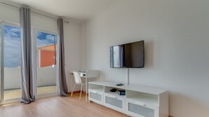 A modern living room with a flat-screen TV mounted on the wall, a white TV stand with storage drawers, a white chair, and a sliding glass door leading to an outdoor area.