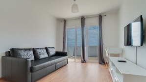 A modern living room with a grey leather sofa, a white desk, and a large window with curtains.