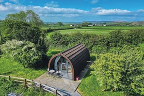 A modern, semi-open structure with a curved roof, a patio area with outdoor seating, and a view of a lush green landscape with distant houses. - Ocean View 6 - 1 Bed Pod - Llanrhidian (Swansea)