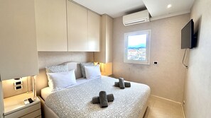 A hotel room with a bed, bedside tables, a TV, and a window with a city view. - Colony (Cannes)