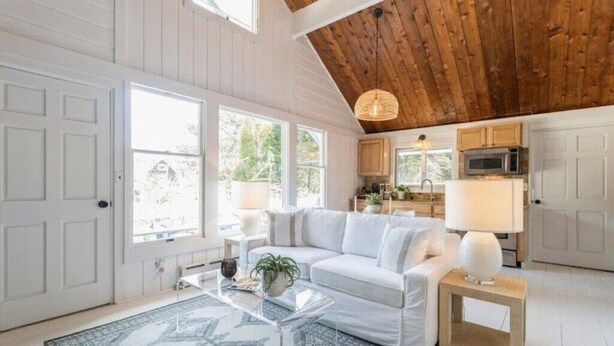 Un vaste espace de vie comprenant un canapé blanc, une table basse et un tapis à motifs. On aperçoit, par une porte ouverte, la cuisine, qui est équipée de meubles en bois et d’un four à micro-ondes.