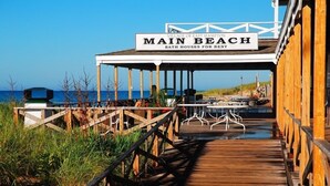 Une promenade en bois mène à une zone abritée, indiquée par une pancarte « Plage principale », et un autre panneau indique « Cabines de plage à louer ».