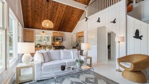 Un vaste espace de vie comprenant un canapé blanc, une table basse et un fauteuil en osier. On aperçoit, au fond, une cuisine avec des meubles en bois et un four à micro-ondes.