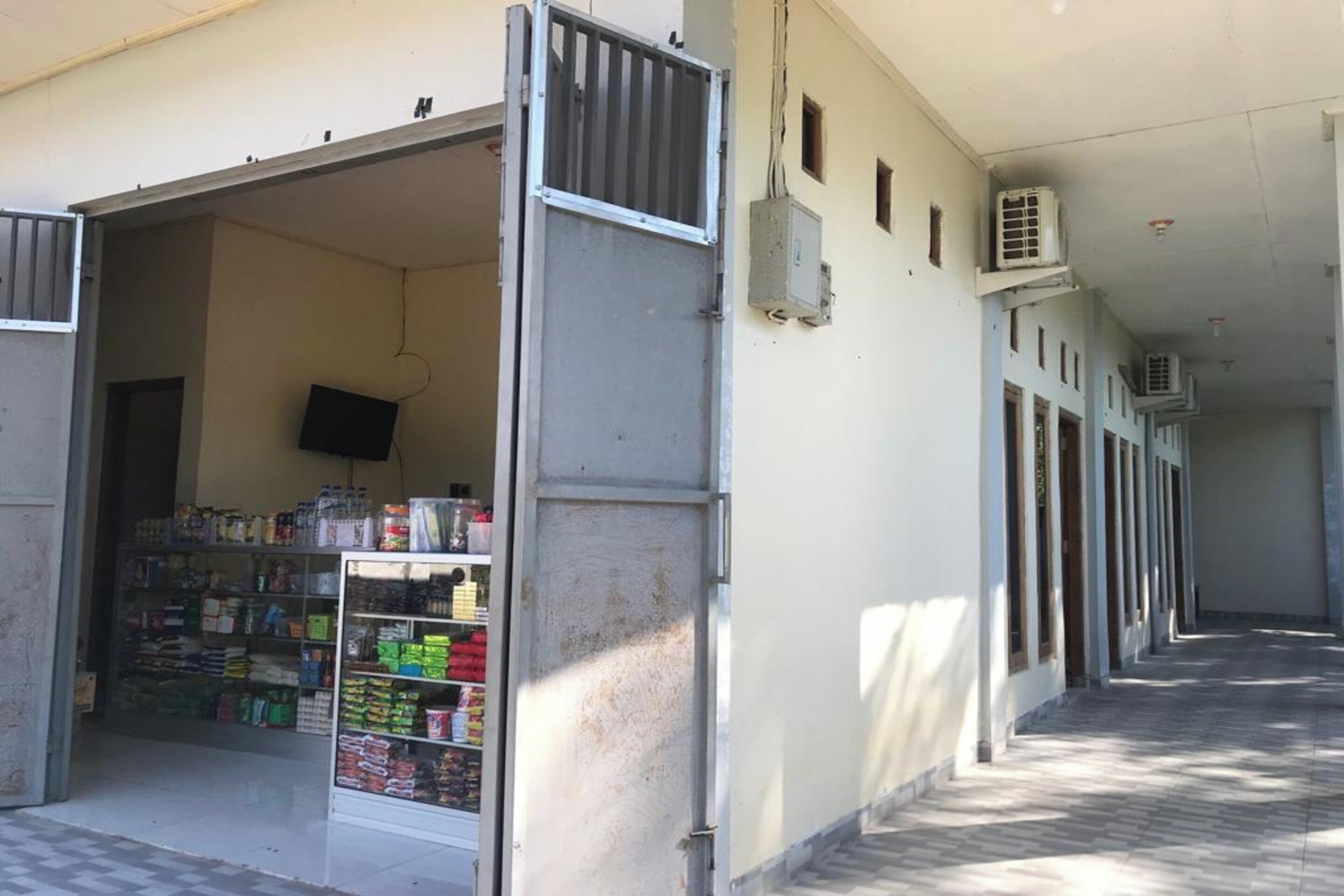 A corridor with a row of air conditioning units on the wall, a closed metal gate, and a store with shelves stocked with various items.