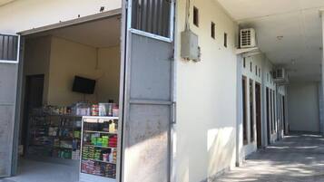 A corridor with a row of air conditioning units on the wall, a closed metal gate, and a store with shelves stocked with various items.