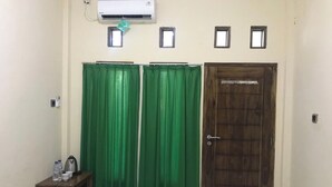 A room with a wooden door, a green curtain, a small table with a kettle and water bottles, and an air conditioning unit on the wall.