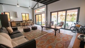 A modern living room with a sofa, coffee table, and ottoman. There is a dining area with a table and chairs, and a kitchen in the background. Large windows provide natural light and views of the outdoors. - Elegant and luxurious apartment in Madrid Tempory (Madrid)