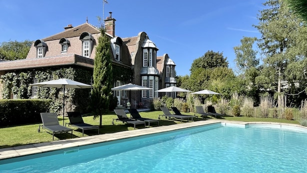 A swimming pool with lounge chairs and umbrellas, surrounded by a well-maintained lawn and a large house with a brick facade and multiple windows. - Maison Héronnière (Compiègne)
