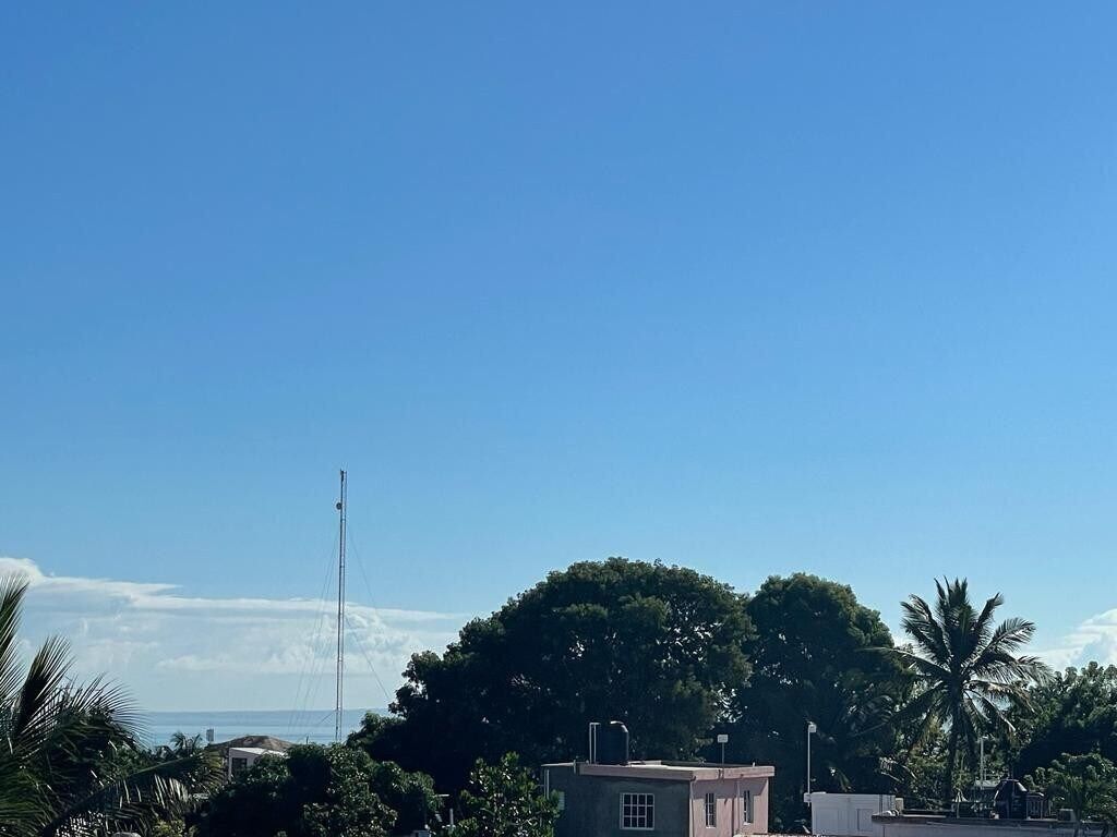 A clear blue sky with a small building and a tall antenna in the foreground, surrounded by lush greenery and palm trees.