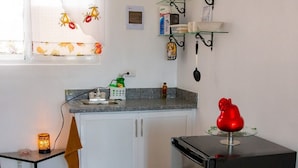 Une cuisine compacte équipée d’un réfrigérateur, d’un évier et d’un petit plan de travail. On y trouve une fenêtre ornée d’un rideau à motifs de fruits, une étagère contenant des ustensiles de cuisine et un objet décoratif rouge posé sur le plan de travail.