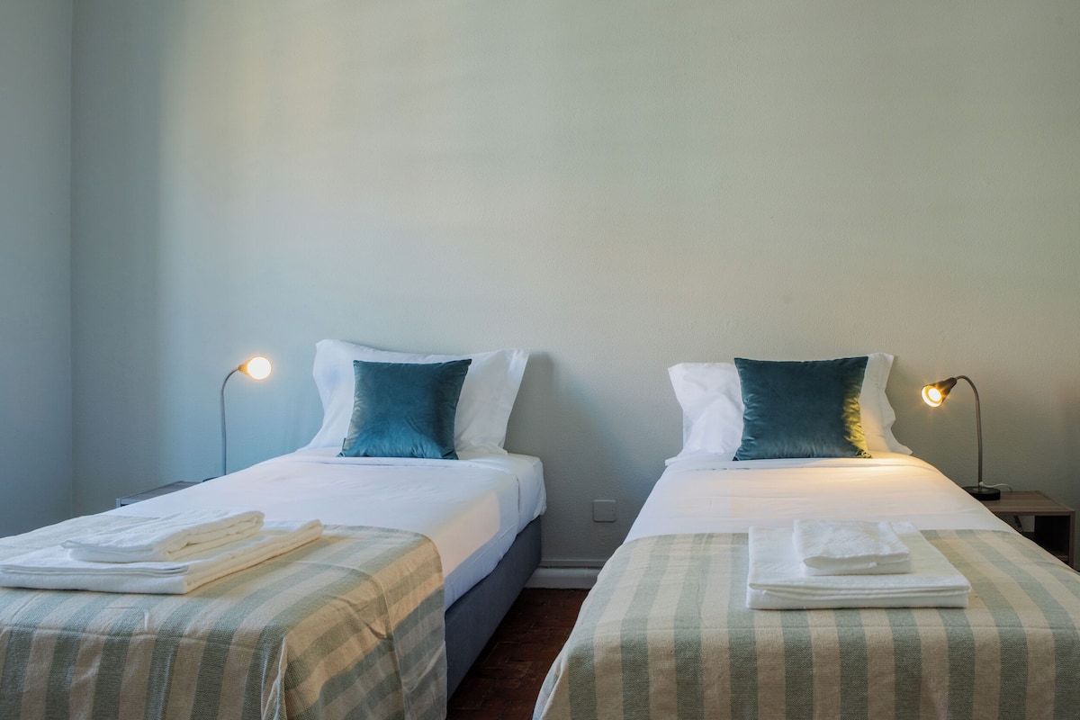 Duas camas de solteiro com lençóis brancos e almofadas azul-turquesa, cada uma com uma toalha dobrada sobre a cama. Há luminárias de cabeceira de cada lado das camas.