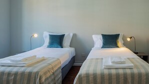Two twin beds with white linens and teal pillows, each with a folded towel on the bed. There are bedside lamps on either side of the beds.