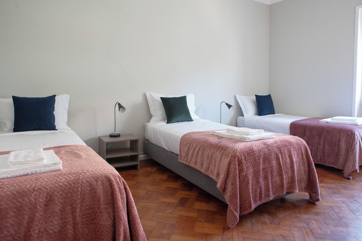 Quarto de hotel com três camas, cada uma com almofada de cor diferente, uma mesa de cabeceira com abajur e piso de madeira.