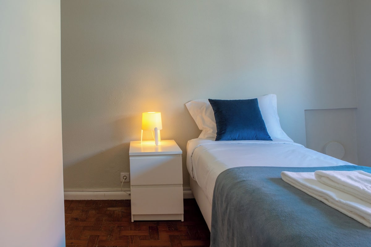 Cama bem arrumada com travesseiro azul e branco, manta cinza e toalha branca dobrada ao lado. Mesa de cabeceira branca com luminária. O quarto tem piso de madeira e paredes em tons claros.