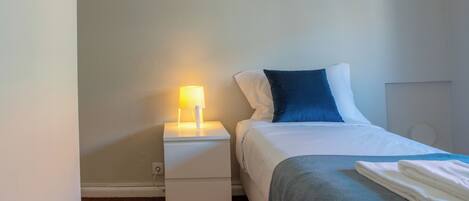 A neatly made bed with a white and blue pillow, a grey blanket, and a folded white towel on the side. There is a white bedside table with a lamp on it. The room has a wooden floor and light-colored walls.