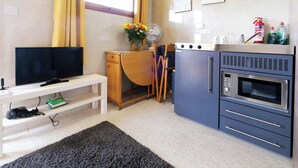 A compact kitchenette with a microwave, oven, and sink. There is a small wooden desk with a chair and a fan. A television is on a white stand, and a rug is on the floor. - Ocean View 7 - 1 Bedroom - Llanrhidian (Swansea)