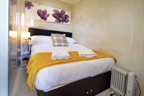 Een net opgemaakt bed met witte lakens, een gele plaid en een ruitkussen. Er staat een radiator aan de muur en een klein tafeltje met een lamp en een plant naast het bed.