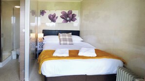 A neatly made bed with white linens and a yellow throw blanket, a bedside lamp, and a wall with floral artwork. - Ocean View 7 - 1 Bedroom - Llanrhidian (Swansea)
