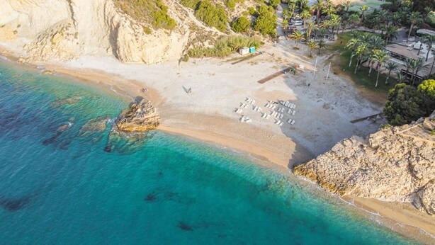 An aerial view of a beach with clear turquoise waters, rocky cliffs, and a small building with a white roof.