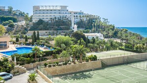 A resort with a swimming pool, tennis court, and a building with multiple balconies overlooking the sea.