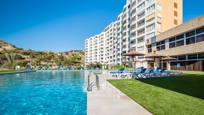 A swimming pool with clear blue water, surrounded by lounge chairs and umbrellas, with a hotel building in the background.