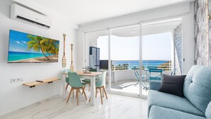 A modern living room with a large TV, a dining area, and a balcony with a view of the ocean.