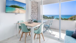 A modern living room with a dining area, a TV mounted on the wall, and a balcony with a view of the sea.