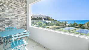A balcony with a view of the sea and tennis courts, a table and chairs, and a textured wall.