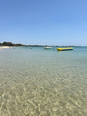En strand med klart, lavt vand, en båd og en fjer borte kystlinje.