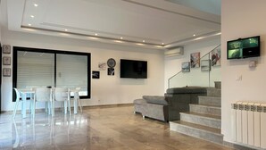A spacious living area with a dining table and chairs, a grey sofa, a flat-screen TV, and a staircase leading to an upper floor. - Villa a Hammamet Centre, 300 Metre du Theatre, Piscine, Climatisée, 4 Chambre (Hammamet)