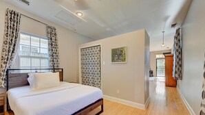 A bedroom with a bed, a window with patterned curtains, a door with a geometric pattern, and a hallway leading to another room. - Cooper Young Vibes Su (Memphis)
