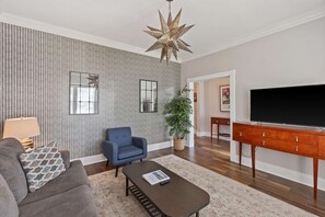 A living room with a grey sofa, a blue armchair, a wooden coffee table, a star-shaped hanging light, a TV stand, and a large plant.