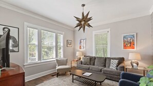 Ein Wohnzimmer mit einer Couch, einem Couchtisch und zwei Lampen. Drei Fenster mit Jalousien, eine sternförmige Leuchte und gerahmte Kunstwerke an den Wänden.