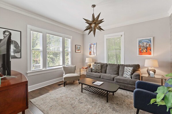 A living room with a sofa, coffee table, and two lamps. There are three windows with blinds, a star-shaped light fixture, and framed artwork on the walls.