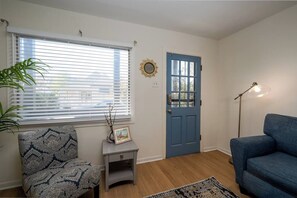 A living room with a blue door, a patterned armchair, a small table with a picture frame, a floor lamp, and a large window with blinds.