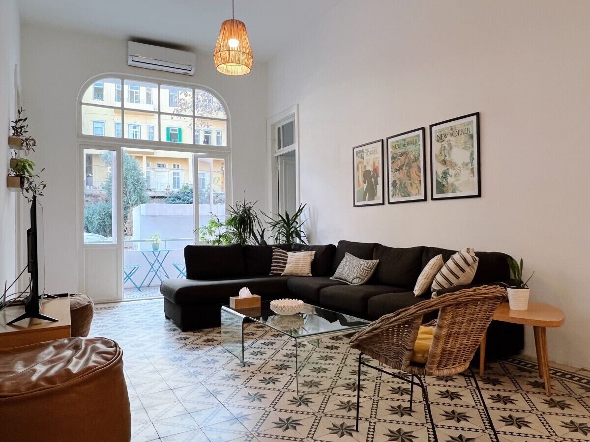 A modern living room with a black sectional sofa, a glass coffee table, and a wicker chair. There are three framed pictures on the wall, a potted plant, and a large window with a view of buildings outside.