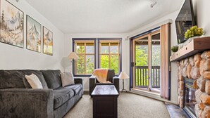 A living room with a gray sofa, a coffee table, and a fireplace. There are three windows and a door leading to an outdoor area.
